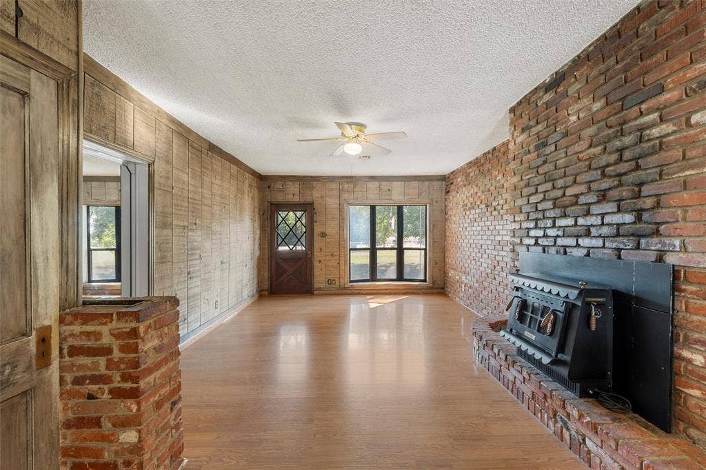 12145 Tree Lake Drive, Waco, TX 76708 Unfurnished living room with a wood stove, wood finished floors, a textured ceiling, ceiling fan, and brick wall