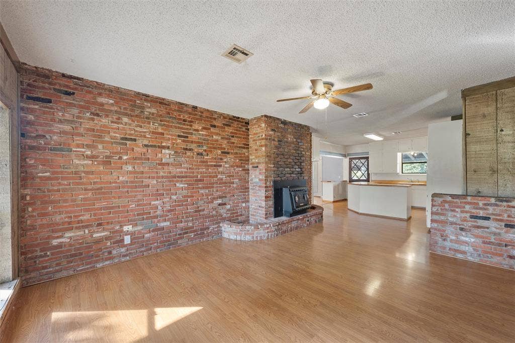 12145 Tree Lake Drive, Waco, TX 76708 Unfurnished living room with brick wall, light wood finished floors, a textured ceiling, a wood stove, and a ceiling fan