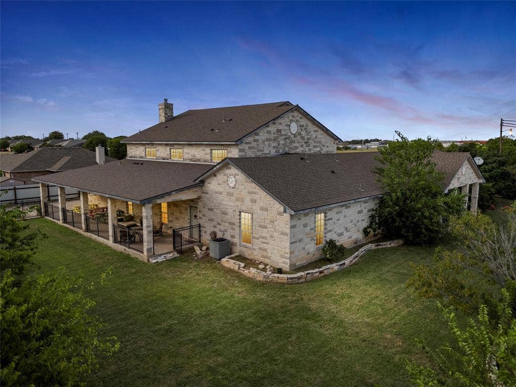 Back of property at dusk with a patio, a chimney, a yard, roof with shingles, and stone siding