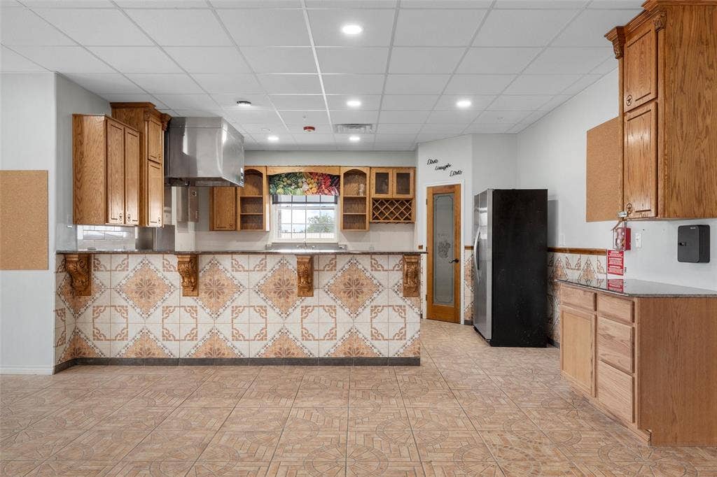 Kitchen featuring glass insert cabinets, a paneled ceiling, freestanding refrigerator, brown cabinetry, and a peninsula