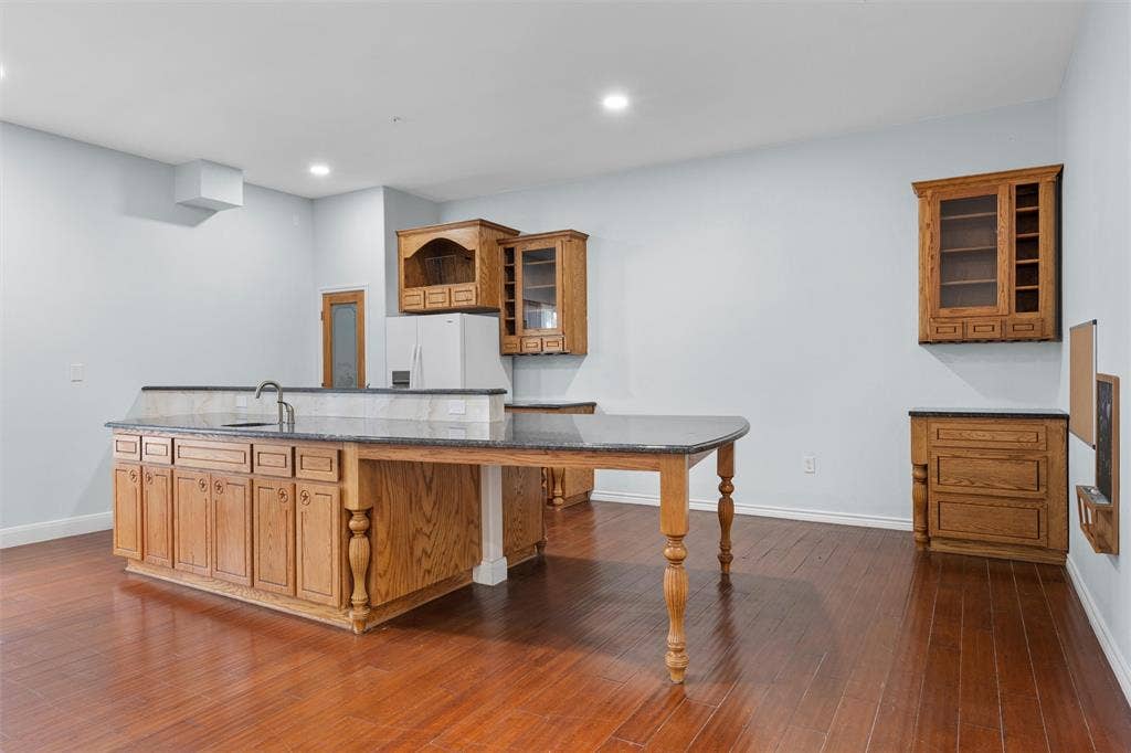 Kitchen featuring brown cabinets, glass insert cabinets, dark stone counters, dark wood-style flooring, and a kitchen island with sink