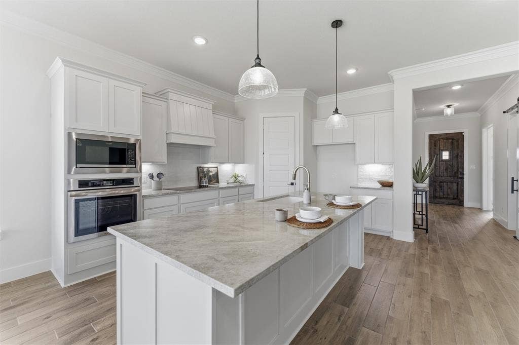 Contemporary kitchen featuring a large central island with a light-toned countertop, white cabinetry with crown molding, stainless steel built-in appliances, and wood-finish flooring