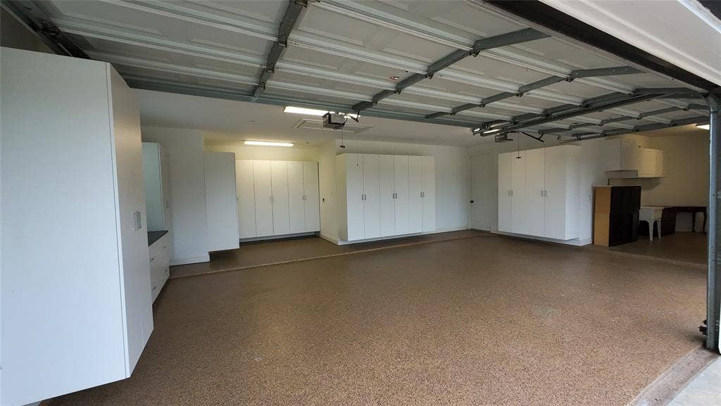 Spacious garage featuring an epoxy-coated concrete floor, extensive built-in white cabinetry, a dedicated utility sink area, and overhead fluorescent lighting