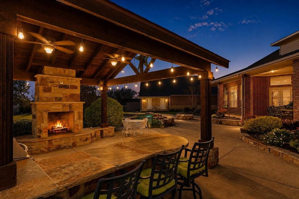Covered outdoor living space featuring a stone fireplace, wood-beamed ceiling, ceiling fans, string lighting, and a built-in stone bar