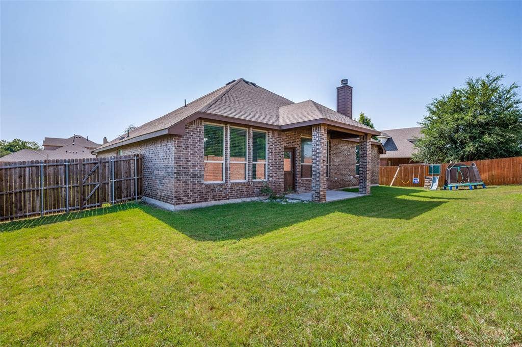 Expansive backyard featuring a manicured lawn, full privacy fencing, brick exterior, covered patio, and a visible chimney