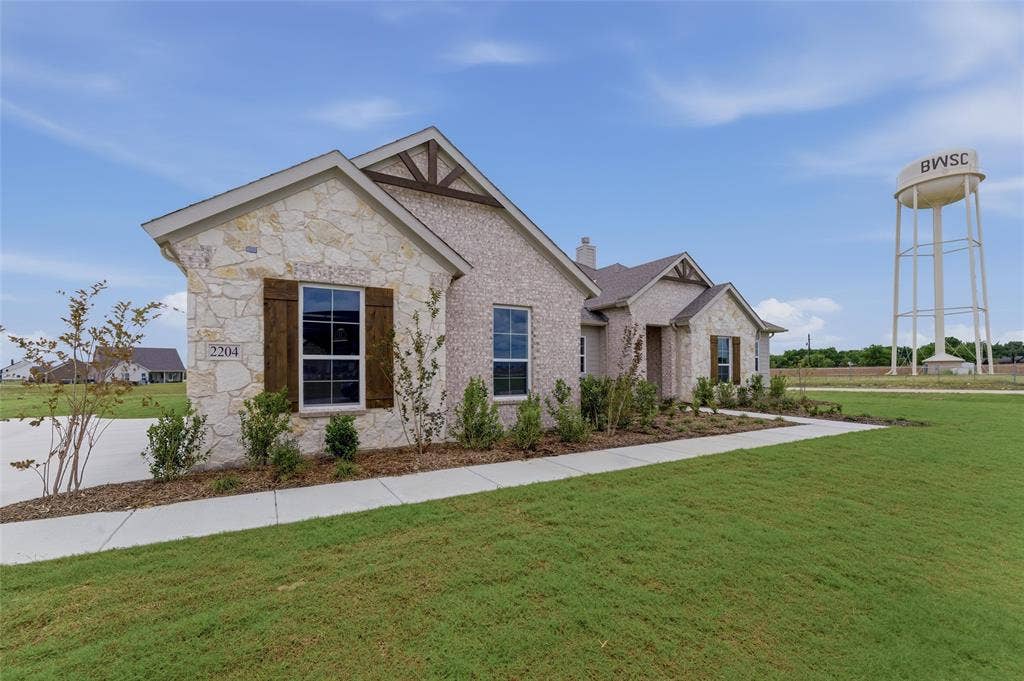 2204 Continent Court, Krum, TX 76249 Stone and brick exterior featuring wooden decorative shutters, a concrete walkway, mature landscaping, and an expansive green lawn