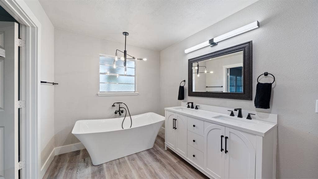 129 King Arthur Road, Mabank, TX 75156 The bathroom features a freestanding tub, a window with privacy glass, a double vanity with white cabinetry and dark hardware, and a dark framed mirror with overhead lighting