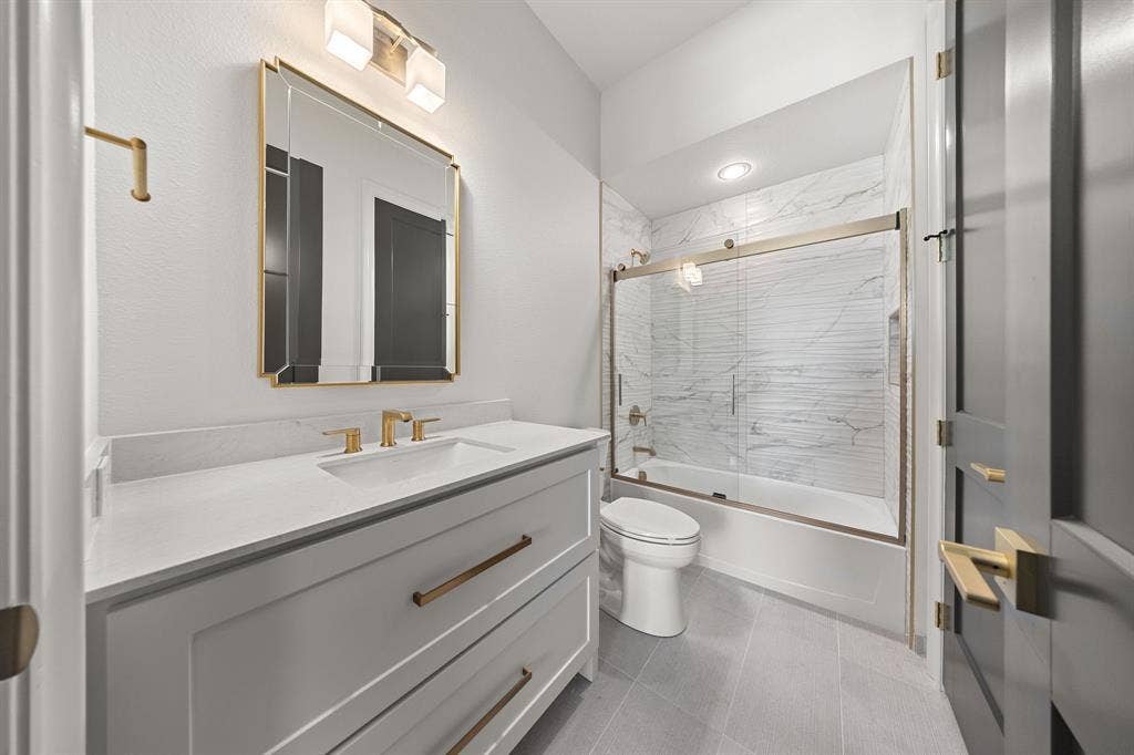 3333 Bridgegate Avenue, Northlake, TX 76247 White shaker vanity with white countertop, gold-finish hardware, gold-finish framed mirror, and a white tiled shower with sliding glass doors