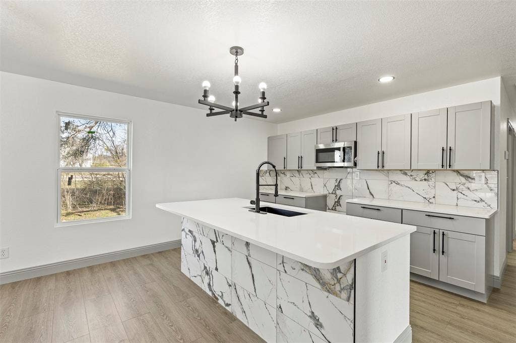 5619 Lake Granbury Trail, Granbury, TX 76048 Kitchen with gray cabinetry, a kitchen island with sink, stainless steel microwave, light wood-style flooring, and light stone countertops
