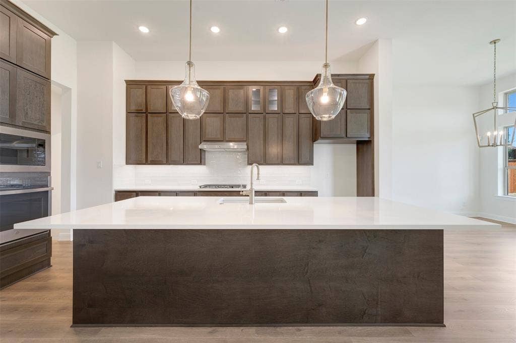 Spacious kitchen featuring a large central island with a white countertop, dark wood-finish cabinetry, built-in stainless steel appliances, and wood-finish flooring