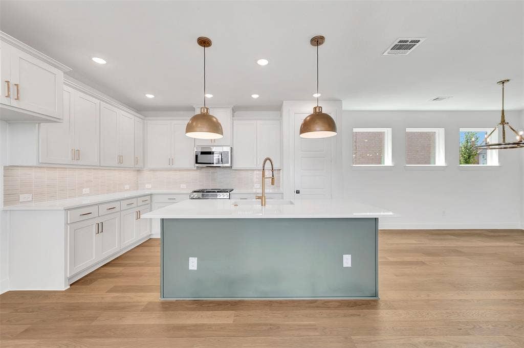 4721 Vizsla Lane, McKinney, TX 75071 Contemporary kitchen featuring white shaker cabinetry, light-toned wood-finish flooring, a central island with a contrasting base and solid surface countertop, stainless steel appliances, and two brass pendant lights