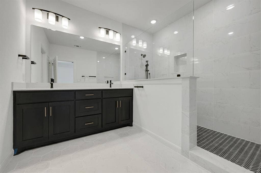 2852 Yampa Drive, McKinney, TX 75071 Contemporary bathroom featuring a dark wood double vanity with white countertops, expansive wall-mounted mirror, and a spacious walk-in shower with white tile walls and a patterned floor