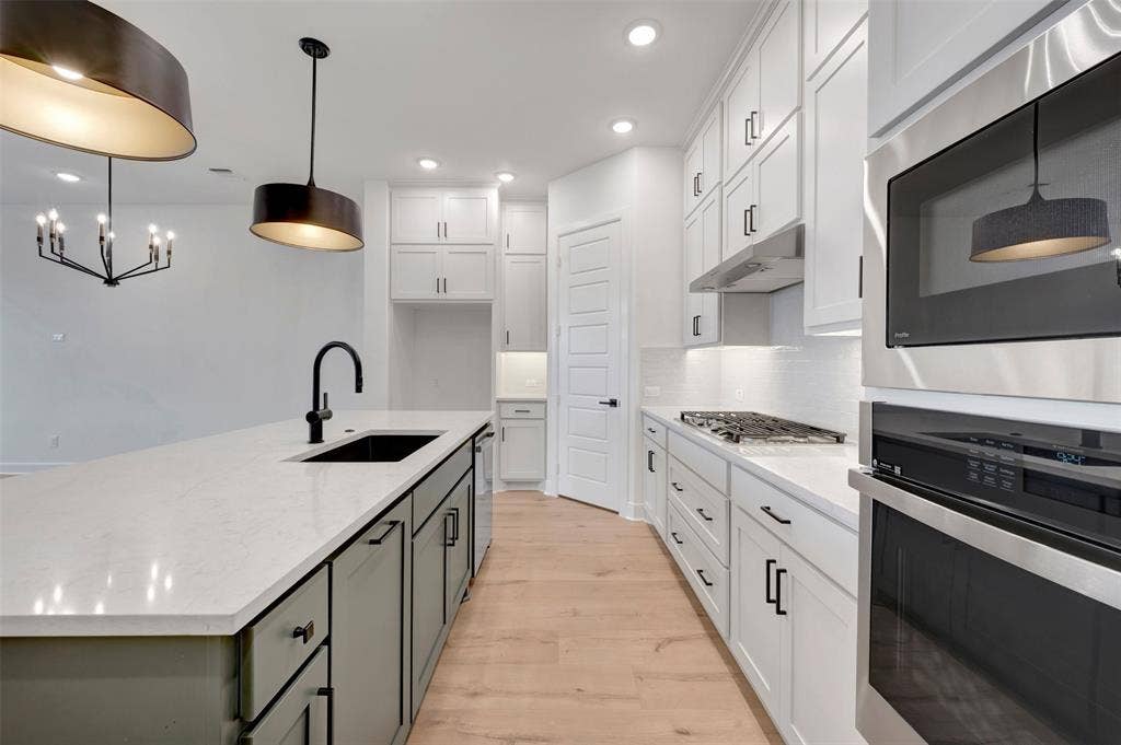 2860 Yampa Drive, McKinney, TX 75071 Contemporary kitchen featuring white shaker cabinetry, stainless steel appliances, a center island with a black sink and matte black faucet, light wood-finish flooring, and pendant lighting fixtures