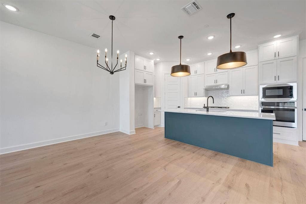 2860 Yampa Drive, McKinney, TX 75071 Open-concept kitchen and dining area featuring light wood-finish flooring, a contemporary black chandelier, a central island with a blue base and white countertop, white shaker cabinetry, and stainless steel appliances