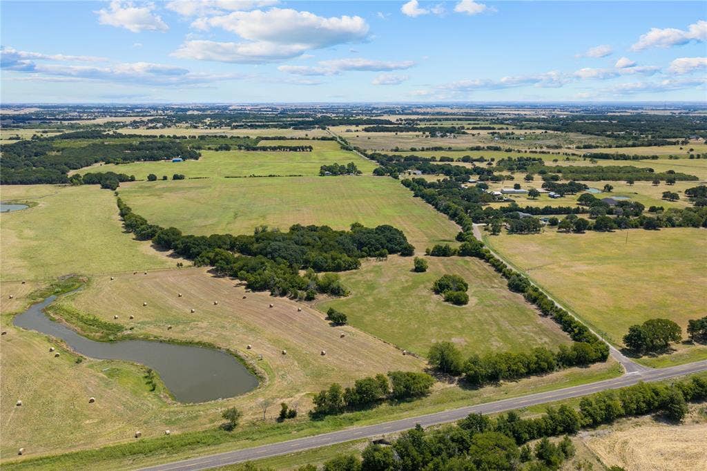 Expansive agricultural land featuring a natural pond, scattered hay bales, mature trees, and a paved road frontage