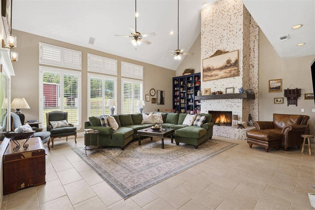 451 Land Of Goshen Drive, Springtown, TX 76082 Vaulted living space featuring a white-washed brick fireplace with a substantial wood mantel, tall windows with plantation shutters, light-toned tile flooring, and built-in shelving