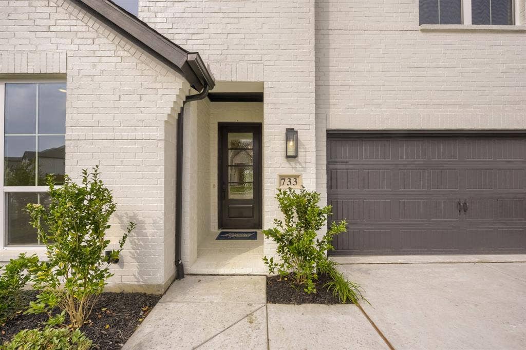 733 Red Cedar Road, Haslet, TX 76052 White brick exterior featuring a dark paneled entry door with glass inserts, a single exterior sconce, and an attached garage with a dark sectional door