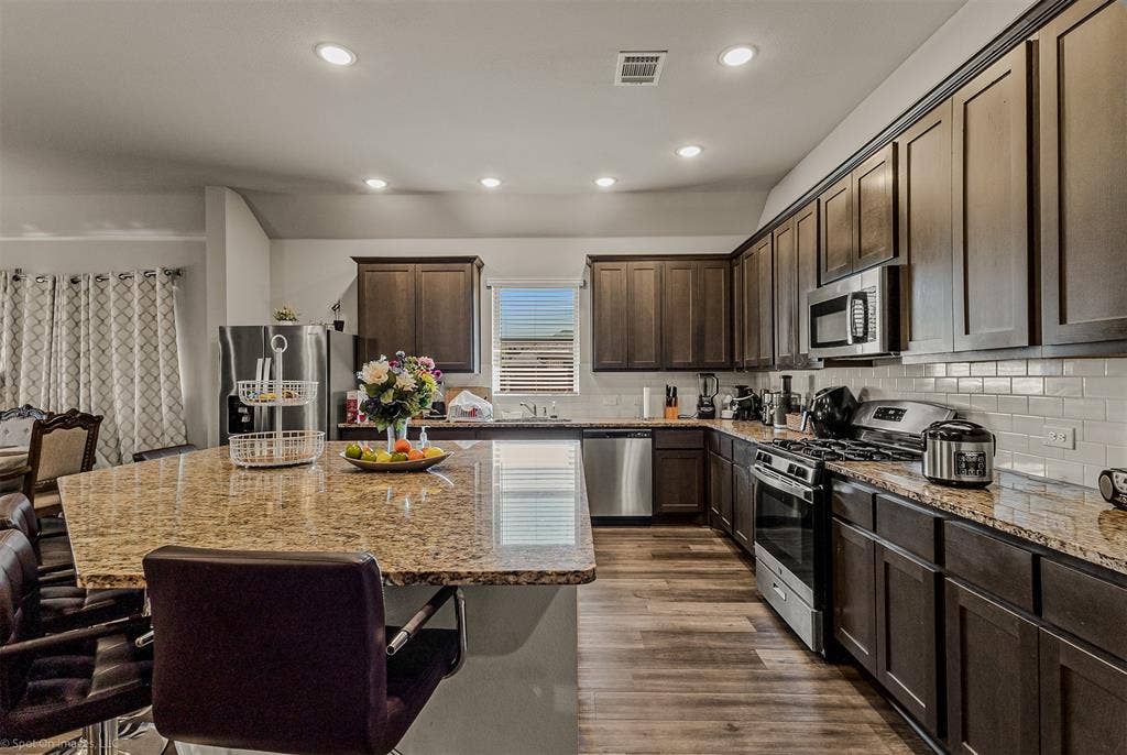5408 Wedge Wood Drive, Haltom City, TX 76137 Kitchen featuring a breakfast bar, dark wood finish cabinets, stainless steel appliances, light stone countertops, and recessed lighting