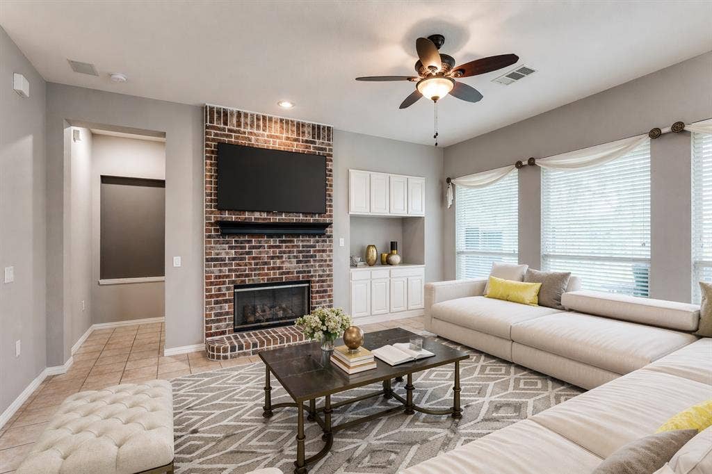 351 Southern Hills Drive, Fairview, TX 75069 Living area featuring a brick fireplace, built-in cabinetry, multiple windows with blinds, a ceiling fan with integrated lighting, and tile flooring - Virtually Staged