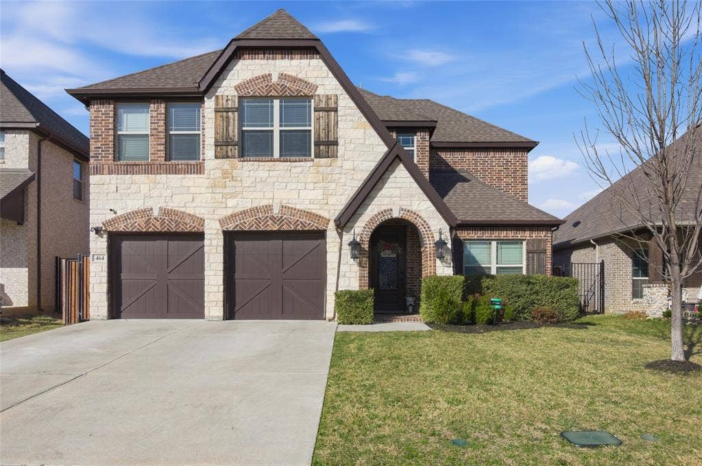 Stone and brick exterior featuring a prominent arched entry, two-car garage with decorative doors, front yard with mature landscaping, and an asphalt shingle roof