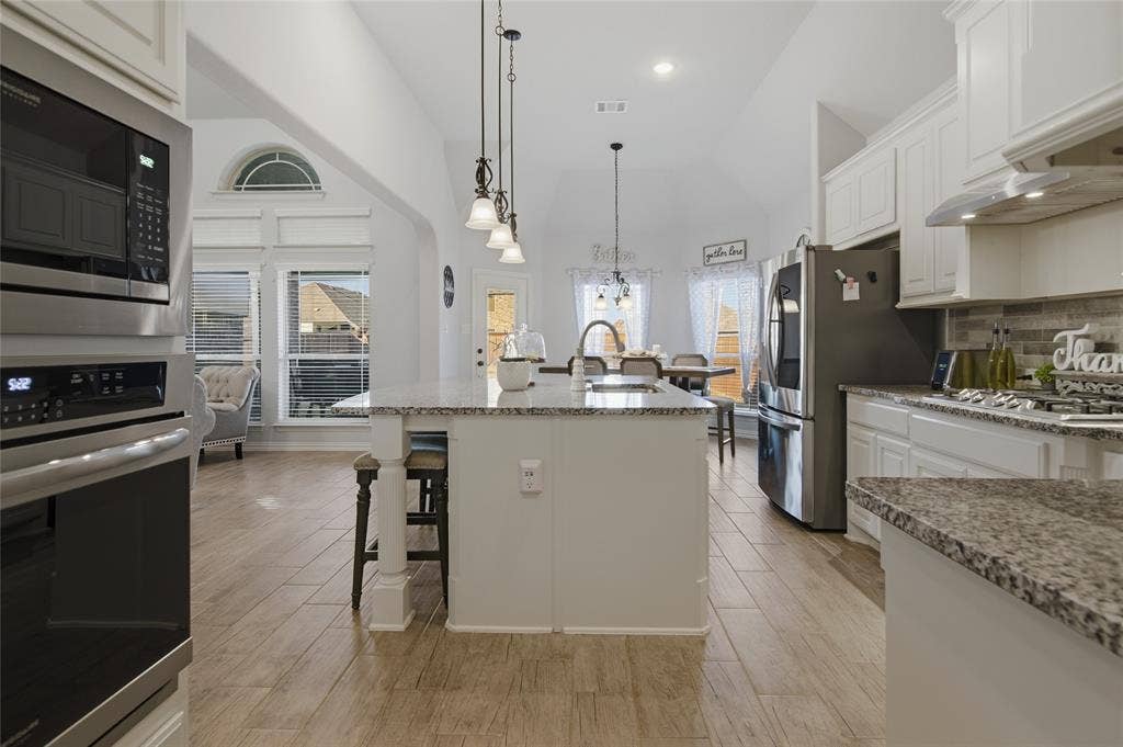 Expansive kitchen featuring a large island with pendant lighting, stainless steel appliances, white cabinetry, granite countertops, and wood-finish flooring