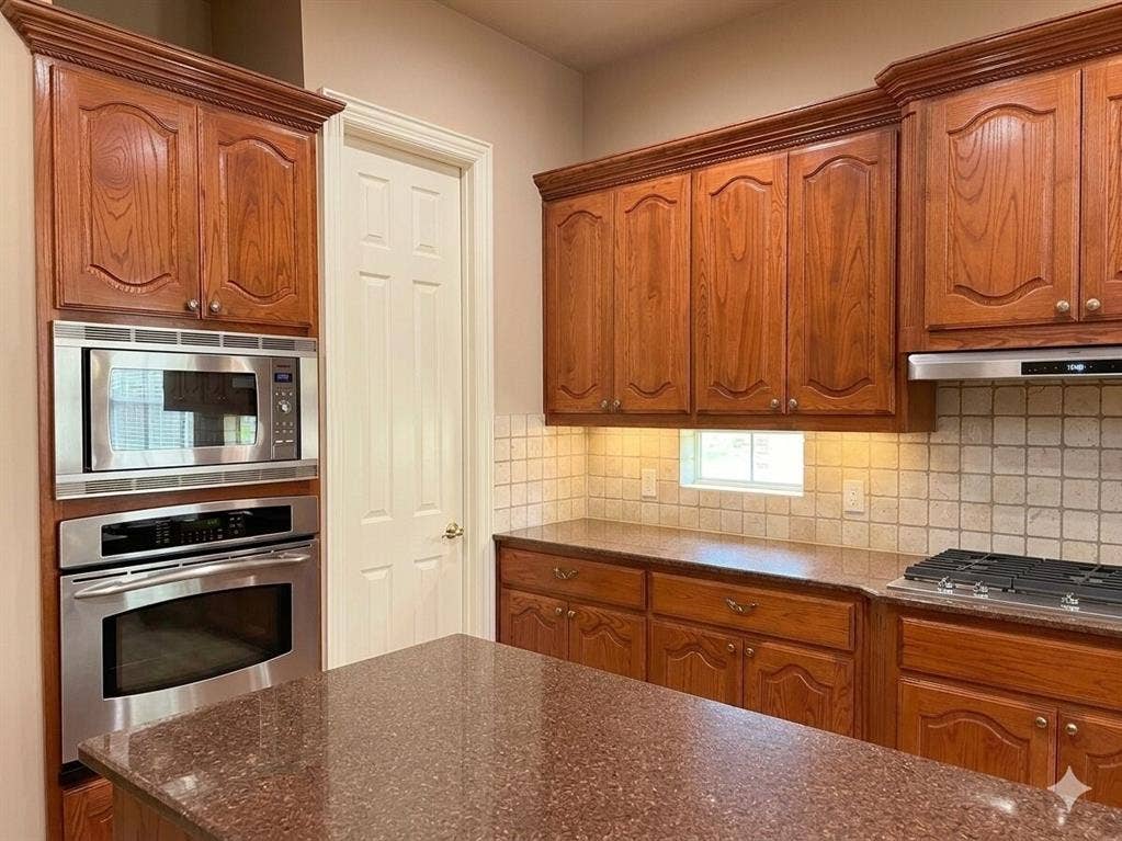 15031 Woodbluff Drive, Frisco, TX 75035 Kitchen featuring rich wood cabinetry with decorative molding, stainless steel built-in microwave and oven, a built-in gas cooktop, tiled backsplash, and stone-finish countertops