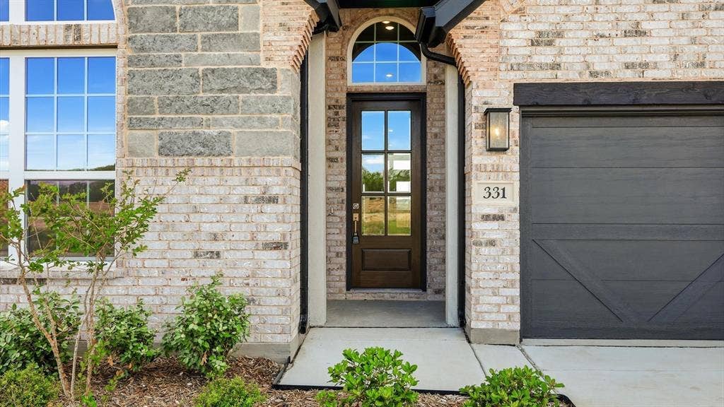 331 Fire Blade Drive, Aledo, TX 76008 Two-tone brick and stone facade featuring an arched entryway with a dark wood-finish door and transom window, a paneled garage door, and modern exterior lighting