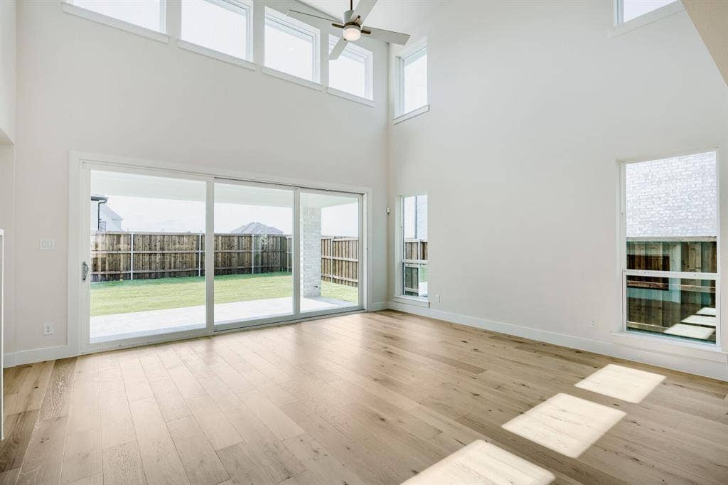940 Heatherbrook Drive, Prosper, TX 75078 Vaulted ceiling great room with wood-finish flooring, featuring multiple high windows, a ceiling fan, and sliding glass doors opening to a private backyard patio and lawn