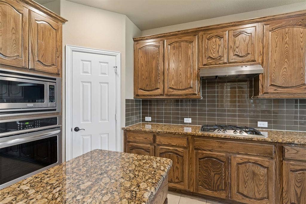 2446 Old Stables Drive, Celina, TX 75009 Kitchen with dark stone countertops, stainless steel appliances, wood finish cabinets, backsplash, and light tile patterned flooring