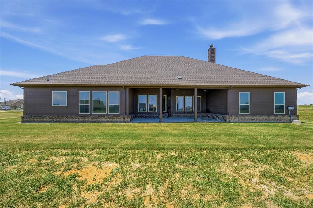 2408 Quartz Street, Krum, TX 76249 Expansive rear exterior featuring a covered patio, multiple large windows, a stone foundation, dark siding, and a shingled roof