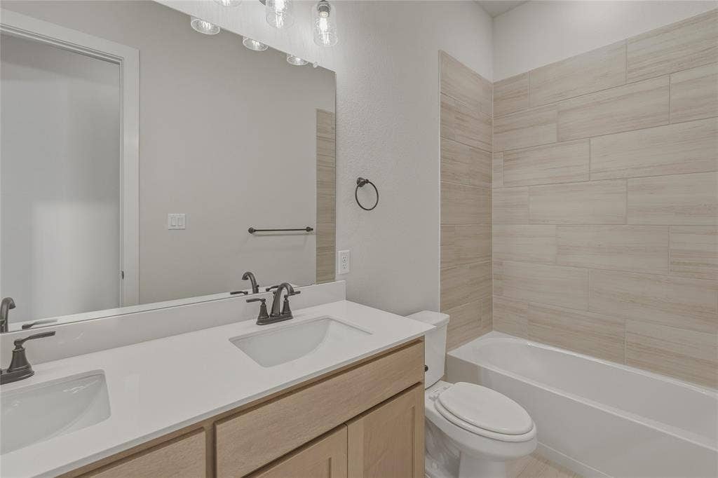 2305 Gypsum Street, Krum, TX 76249 Bathroom featuring a double vanity with light wood-finish cabinetry and white countertops, dual undermount sinks with bronze-finish fixtures, a large frameless mirror, and a tiled shower-tub combination