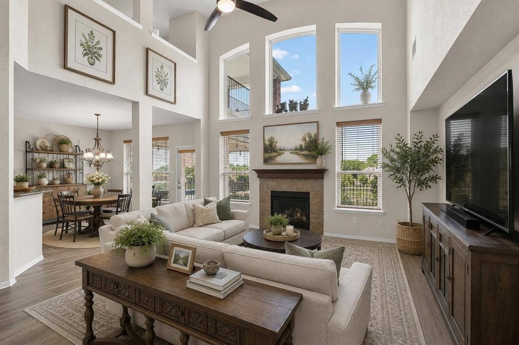 5212 Whisper Drive, Fort Worth, TX 76123 Virtually Staged Two-story living room featuring a fireplace with a stone surround, multiple tall windows, a ceiling fan, and wood-finish flooring