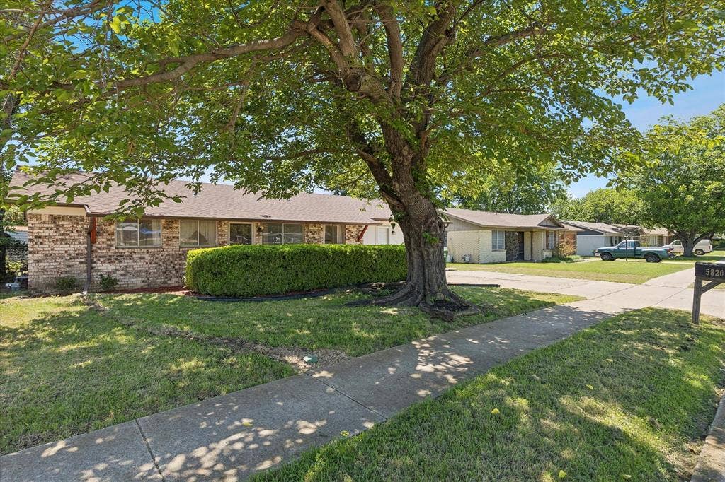 5820 Shipp Drive, Watauga, TX 76148 Brick facade property with a mature shade tree, a manicured lawn, and a paved sidewalk