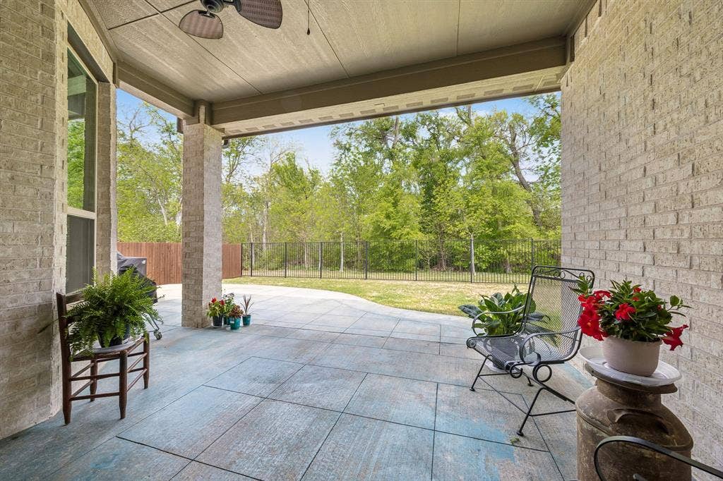 Expansive covered outdoor patio that transitions into a spacious grassy yard framed by a black wrought iron fence along the perimeter. A beautiful line of mature, dense green trees lines the back of the property offering a scenic and private natural views.