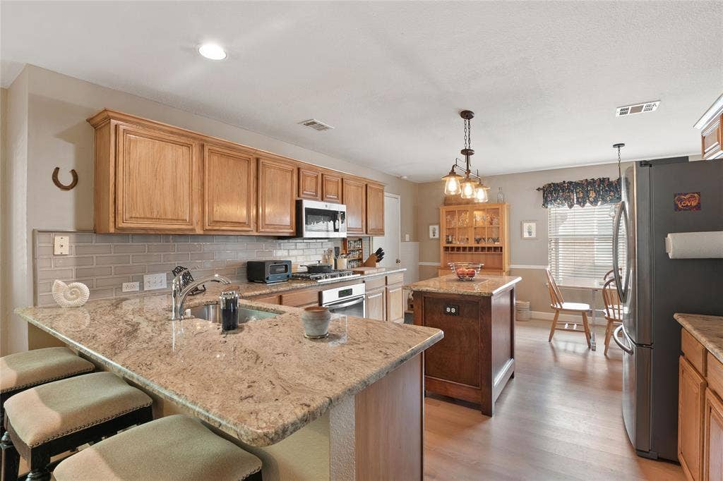 1608 Mountain Laurel Drive, Keller, TX 76248 Kitchen featuring an island with a pendant light fixture, granite countertops, light wood-finish cabinetry, a subway tile backsplash, and stainless steel appliances