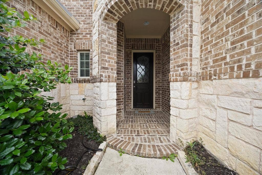 Arched entryway featuring a dark wood-finish door with decorative glass, brick and stone facade, and a brick paver porch