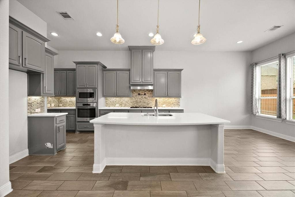 Spacious kitchen featuring an extended center island with integrated sink, sleek grey cabinetry, built-in stainless steel appliances, a mosaic tile backsplash, and wood-finish flooring