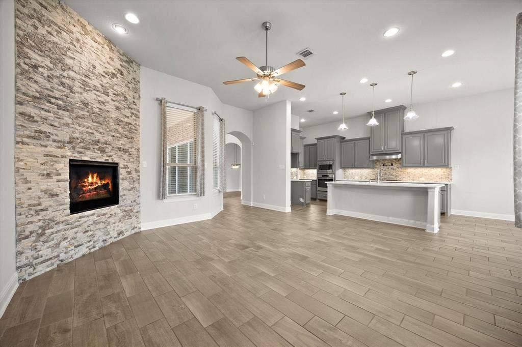 Stone-clad fireplace feature wall, wood-finish flooring, recessed lighting, ceiling fan, and a kitchen with gray cabinetry and pendant lights