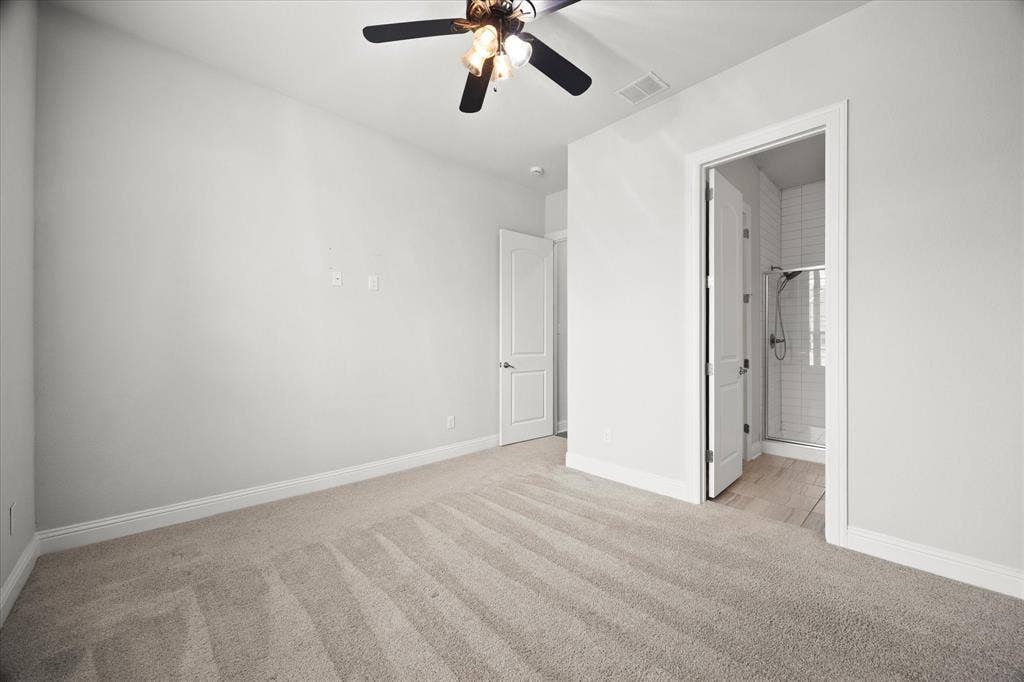 Neutral tone carpeted room featuring a ceiling fan, baseboard molding, a white interior door, and an entryway to a bathroom with a glass-enclosed shower and subway tile surround