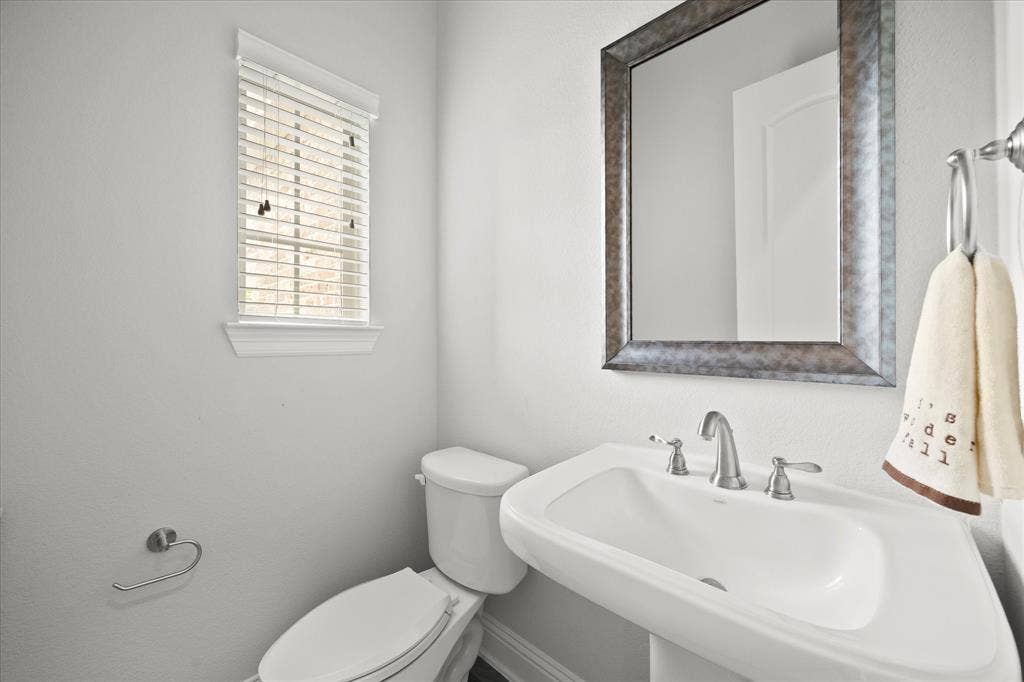Pedestal sink with a brushed nickel faucet, a rectangular framed mirror, a privacy window with blinds, and white toilet