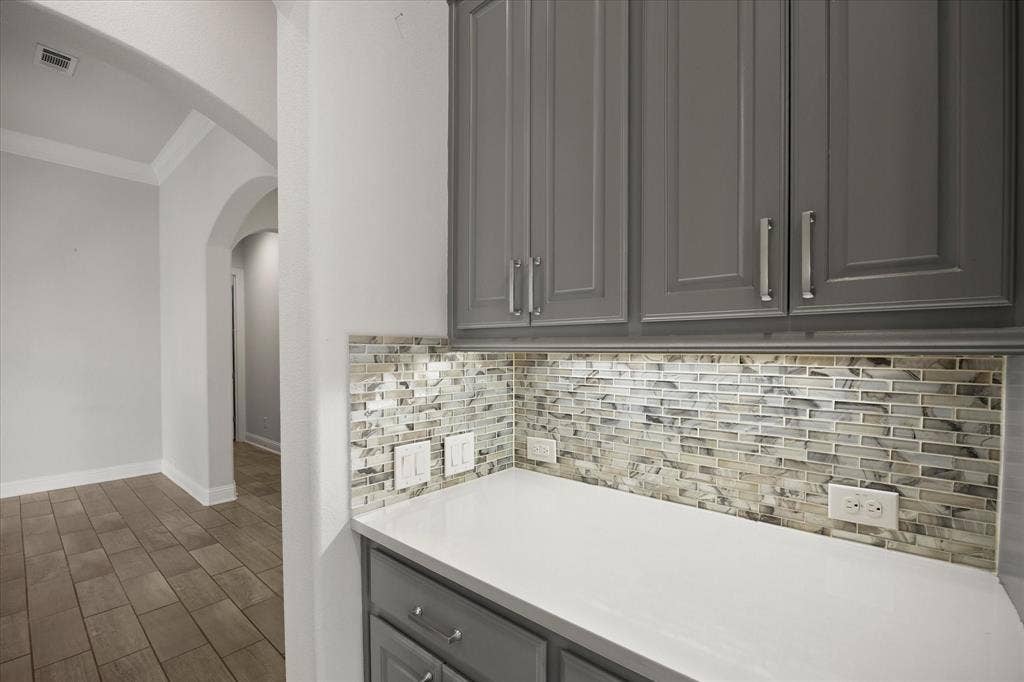 Gray cabinetry with polished chrome hardware, a white countertop, and a mosaic tile backsplash with under-cabinet lighting