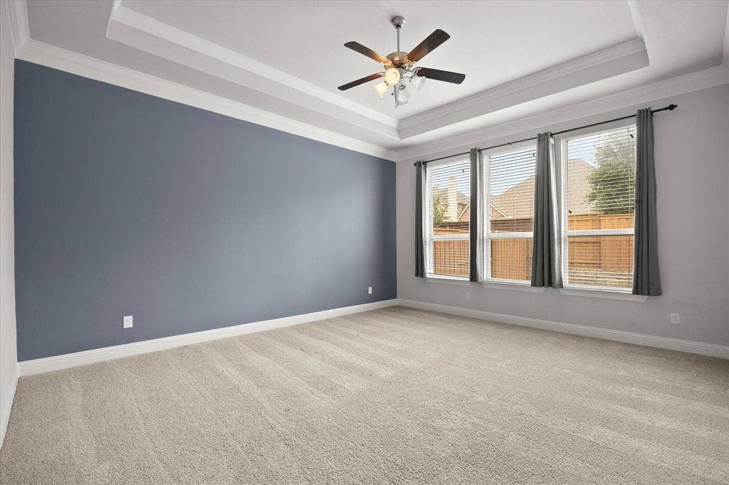 Expansive room featuring tray ceiling, crown molding, a ceiling fan with integrated lighting, three large windows with blinds, and soft carpeting