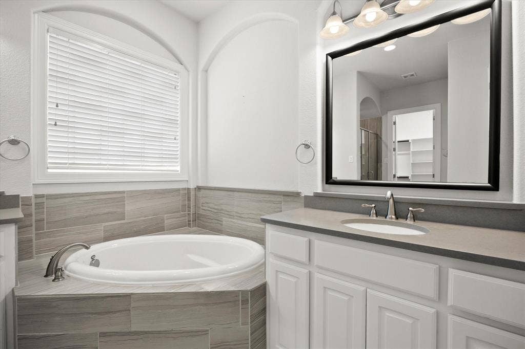 Bathroom featuring a built-in soaking tub with tile surround, a single vanity with a gray countertop, and a large framed mirror with overhead lighting