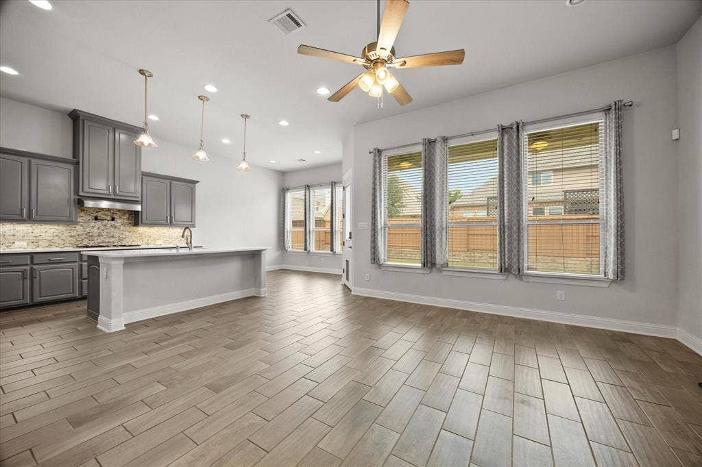 Spacious open-concept living area featuring wood-finish flooring, a ceiling fan with integrated lighting, and multiple windows with blinds and decorative drapes