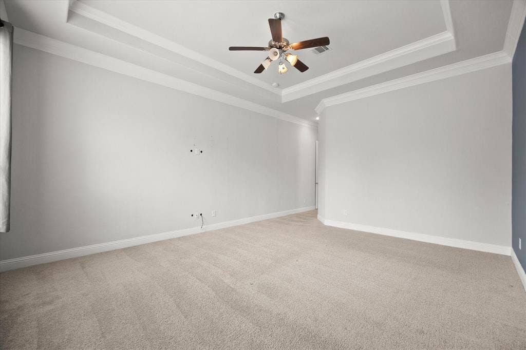Spacious carpeted room featuring a tray ceiling with crown molding, a ceiling fan with integrated lighting, and neutral wall tones