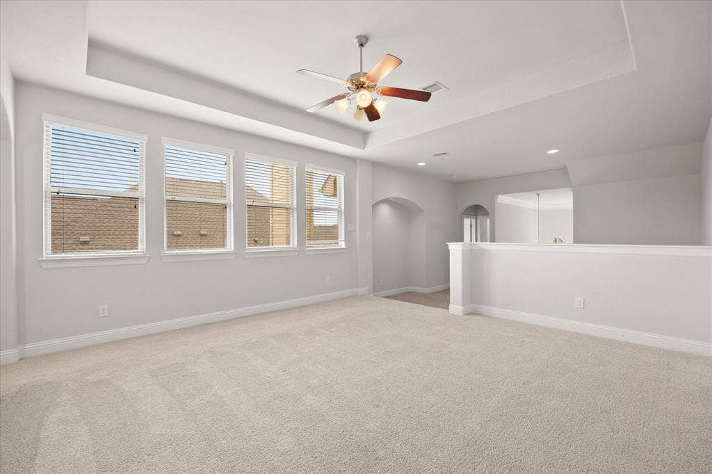 Spacious room featuring light-colored carpeting, a tray ceiling with a ceiling fan, and multiple windows with blinds