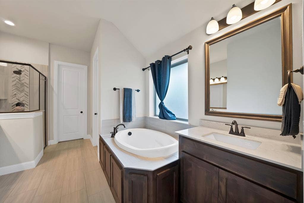 1905 Silver Falls Drive, Burleson, TX 76028 The bathroom features a built-in tub, a dark wood vanity with a white countertop and rectangular sink, and a glass-enclosed shower with chevron-patterned tiling