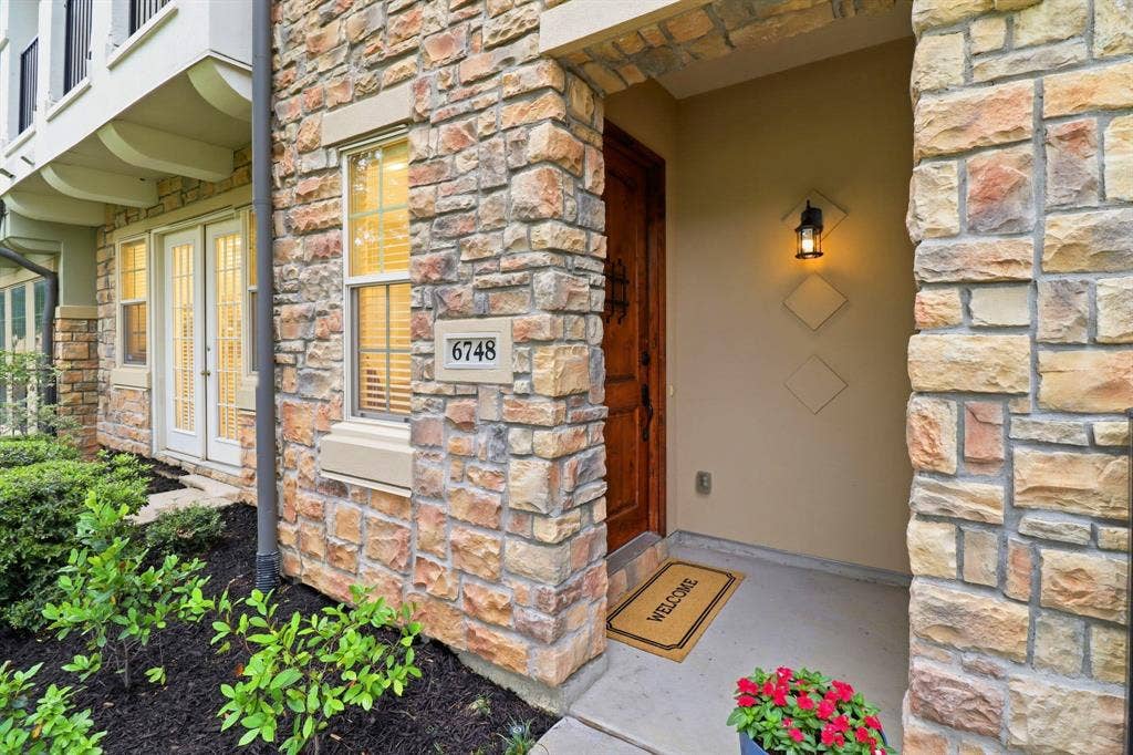 6748 San Roque, Irving, TX 75039 The property features an exterior facade of varied natural stone, a wooden entry door, and an exterior light fixture