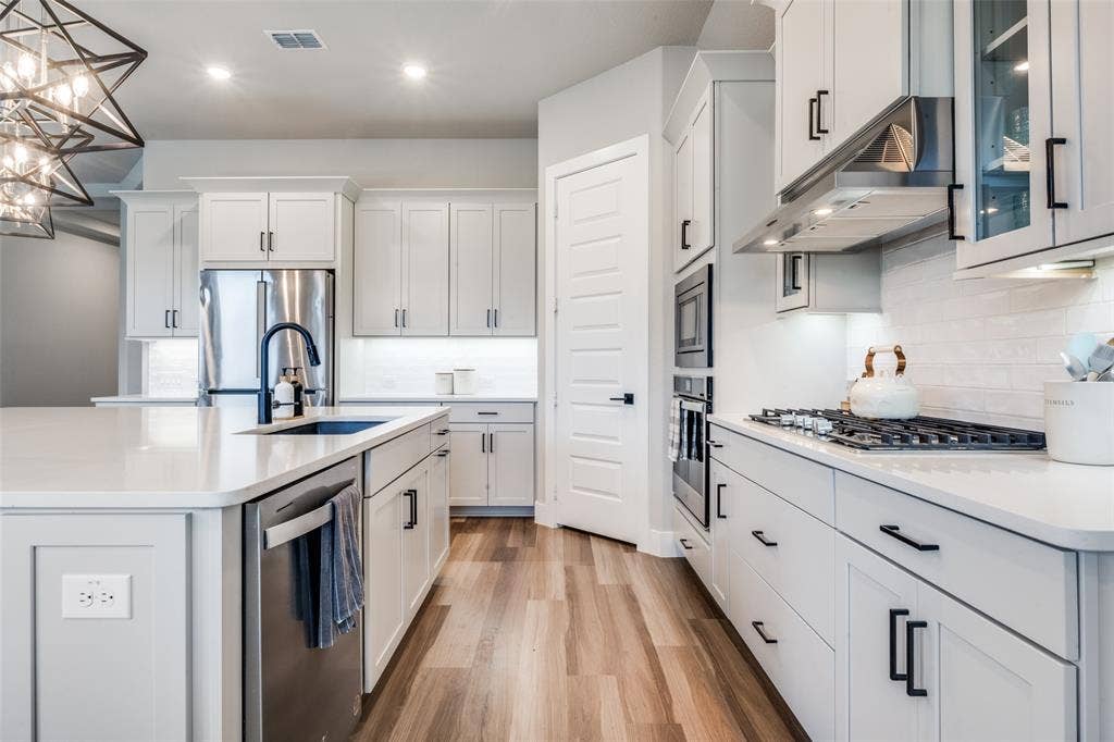 2801 Bedford Road, Northlake, TX 76226 Modern kitchen featuring a large central island, farmhouse sink, stainless steel appliances, sleek white cabinetry with contemporary black hardware, and tiled backsplash. Perfectly arranged for cooking and entertaining.
