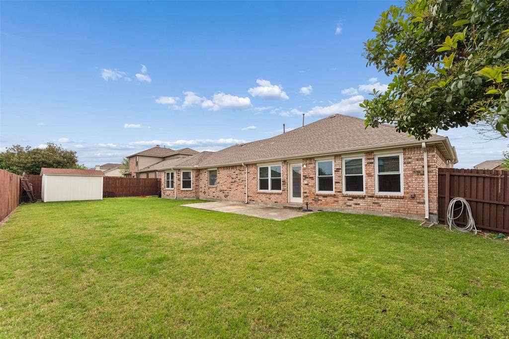 1221 Amistad Drive, Prosper, TX 75078 The property features a brick exterior, a well-maintained lawn, a privacy fence, and a patio area